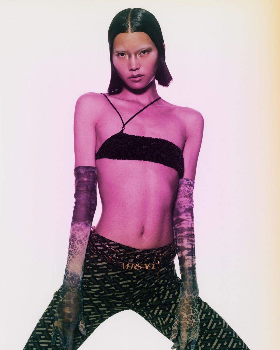 A striking fashion portrait features an Asian model wearing a black top, patterned pants, and sheer gloves against a soft pink backdrop.