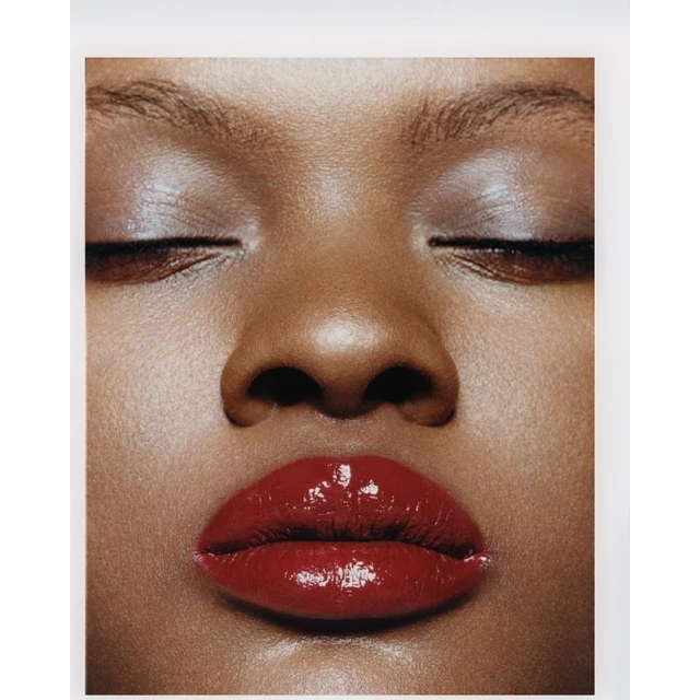 Close-up portrait of an adult female with closed eyes and glossy red lipstick. High-key lighting emphasizes the skin's texture and tone.