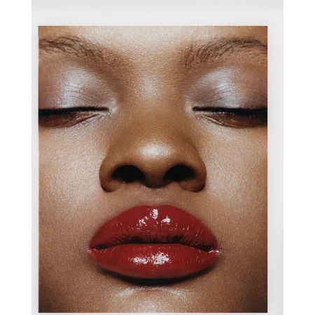 Close-up portrait of an adult female with closed eyes and glossy red lipstick. High-key lighting emphasizes the skin's texture and tone.