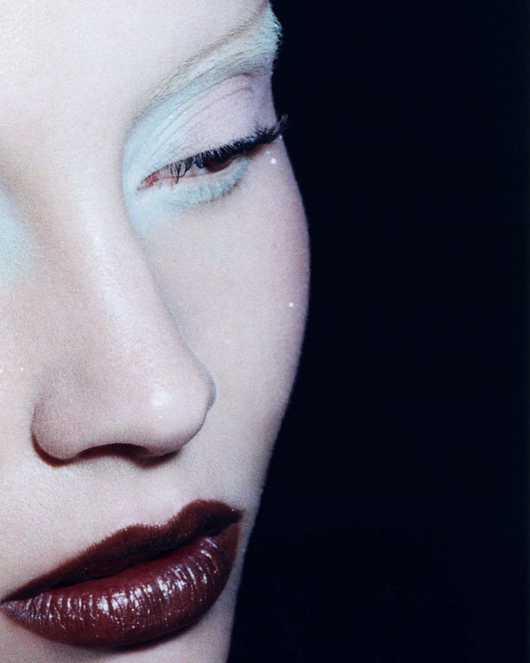 Close-up portrait of a person with fair skin, red lips, and light-colored eyebrows and eyeshadow, set against a dark black background.