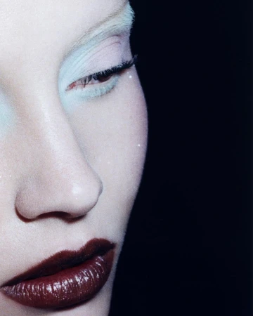Close-up portrait of a person with fair skin, red lips, and light-colored eyebrows and eyeshadow, set against a dark black background.