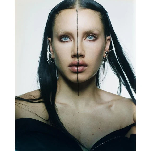 Close-up portrait of a young woman with fair skin, dark hair, and light blue eyes. A vertical line bisects her face, and she wears jewelry.