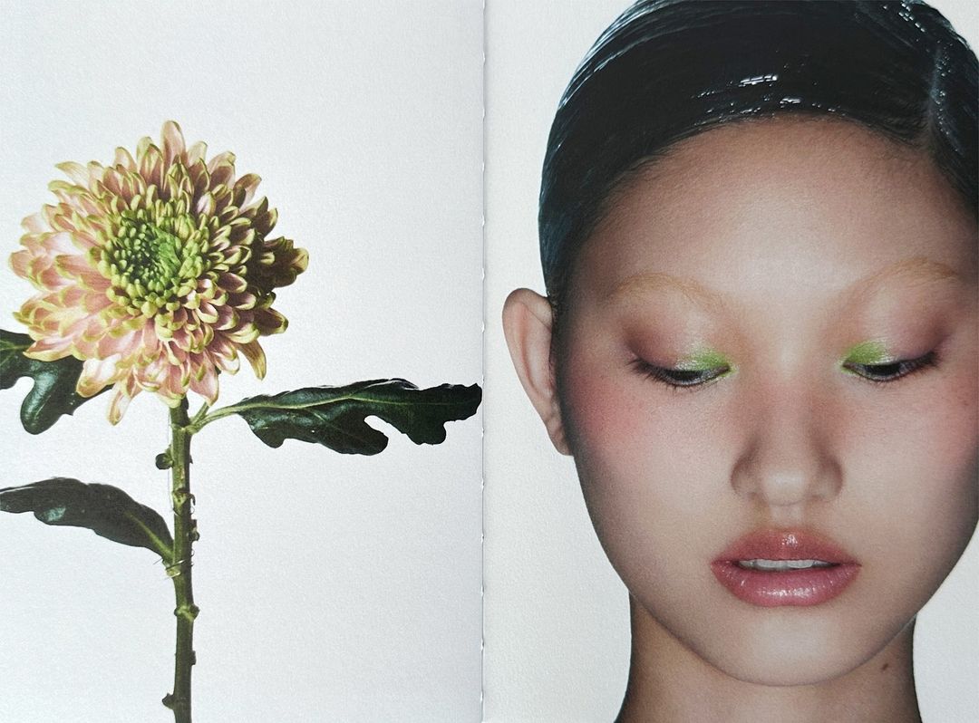 Close-up studio shot of an Asian woman with wet-look hair and green eyeshadow next to a blooming dahlia flower on a white backdrop.