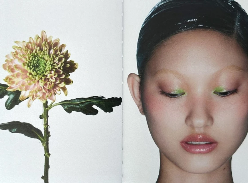Close-up studio shot of an Asian woman with wet-look hair and green eyeshadow next to a blooming dahlia flower on a white backdrop.