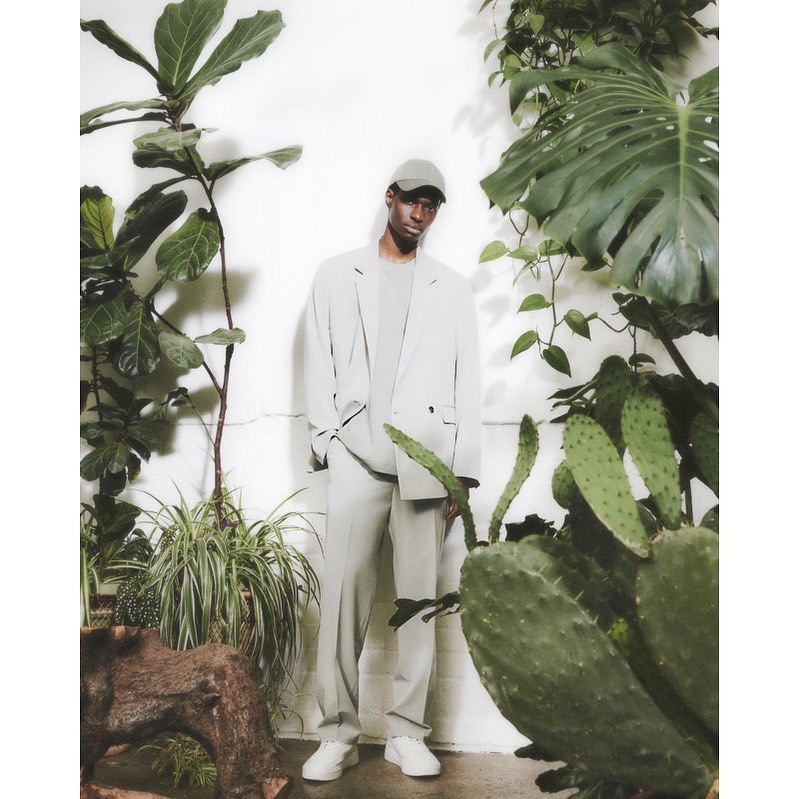 A Black male model poses in a light gray suit surrounded by lush green plants against a white wall in a studio setting.