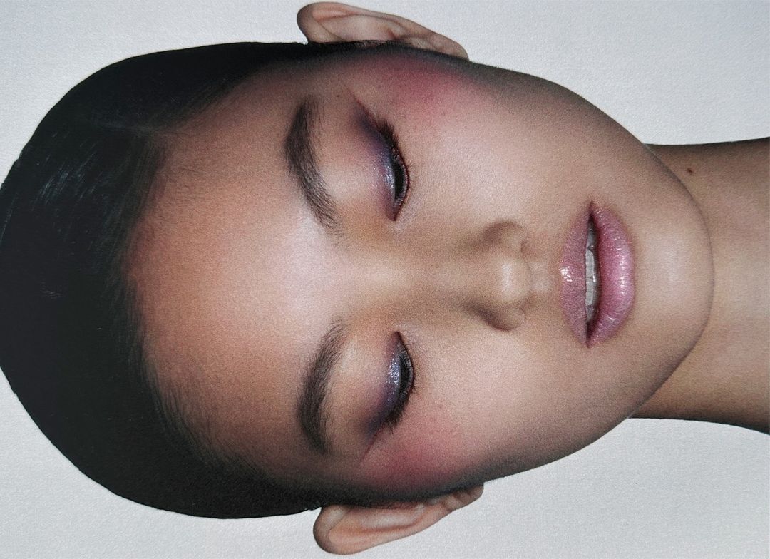 Close-up beauty portrait of a woman with closed eyes, soft purple eyeshadow, and glossy pink lips against a plain backdrop.