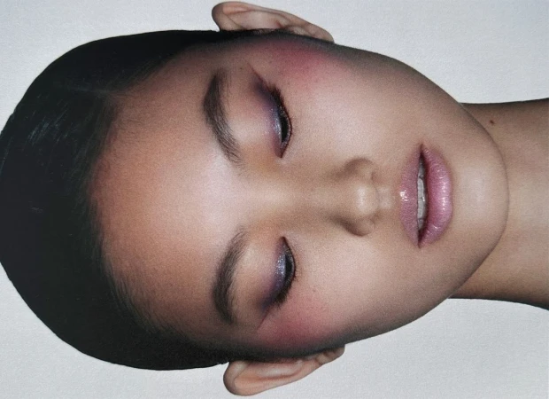 Close-up beauty portrait of a woman with closed eyes, soft purple eyeshadow, and glossy pink lips against a plain backdrop.
