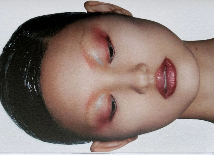 Close-up portrait of a woman with glossy black hair and shimmery makeup featuring red eyeshadow and lipstick, viewed from an angle against a white background.