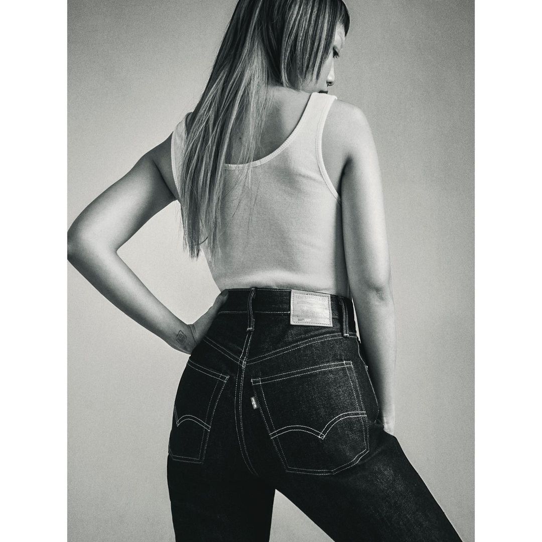Monochrome portrait of a woman wearing a white tank top and dark high-waisted jeans against a plain studio background.