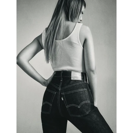 Monochrome portrait of a woman wearing a white tank top and dark high-waisted jeans against a plain studio background.