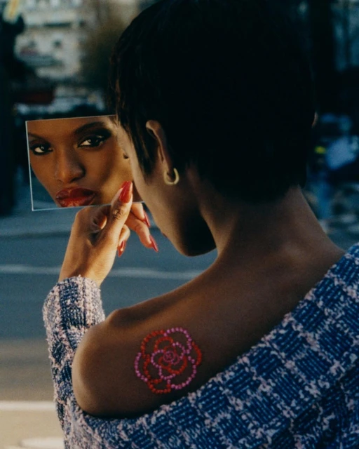 Woman views her reflection in a mirror outdoors, showcasing red lipstick, a patterned jacket, and a rose tattoo on her shoulder.