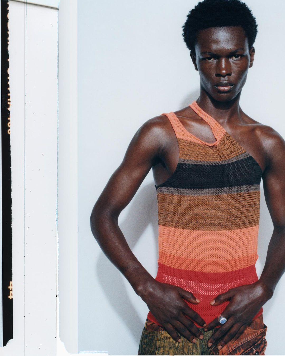 Portrait of a Black male model wearing a brown and orange striped one-shoulder top with hands near waist against white wall indoors.