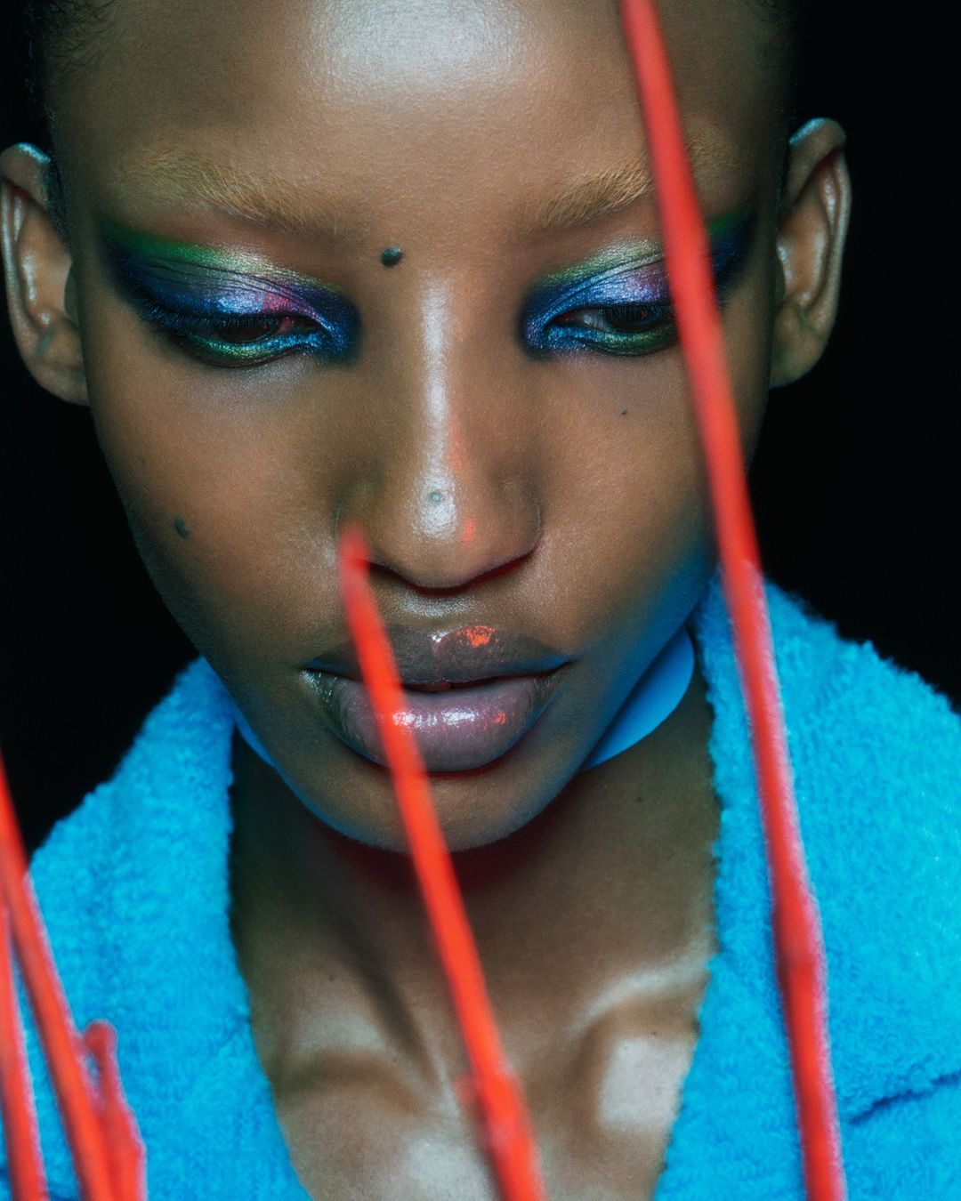 Close-up portrait of a Black woman wearing blue textured apparel with vibrant eyeshadow and red elements in the foreground.