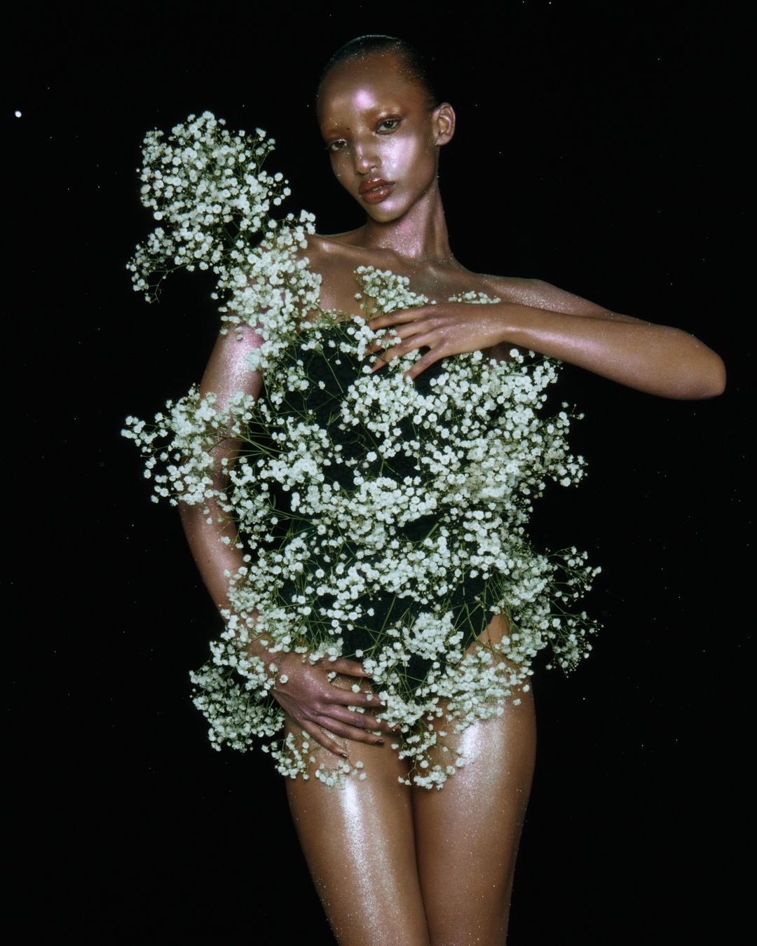Artistic portrait of a model covered in white baby's breath flowers against a black background in a studio setting, shimmering skin.