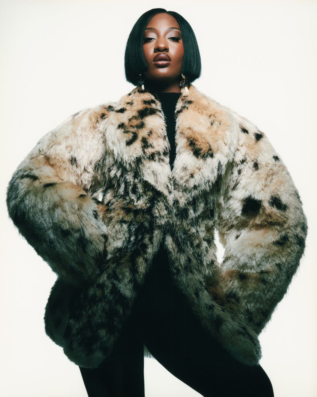 Portrait of an adult woman wearing a large faux fur coat in a studio setting, looking directly at the camera with neutral expression.
