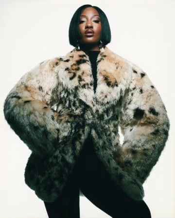 Portrait of an adult woman wearing a large faux fur coat in a studio setting, looking directly at the camera with neutral expression.