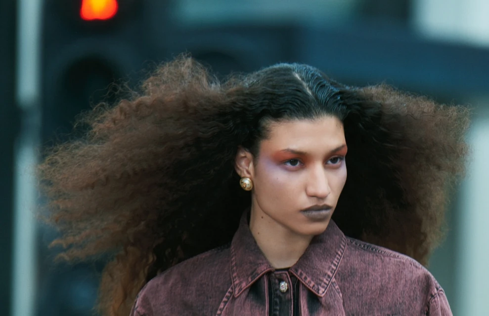 Close-up of a fashion model with curly hair and dramatic makeup, wearing a red denim jacket against a blurred urban background.