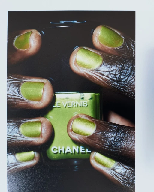 Close-up of dark-skinned hands with metallic green nail polish holding Chanel nail polish bottle with same color in a dark setting.