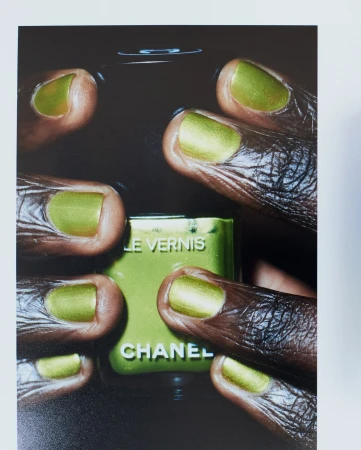 Close-up of dark-skinned hands with metallic green nail polish holding Chanel nail polish bottle with same color in a dark setting.
