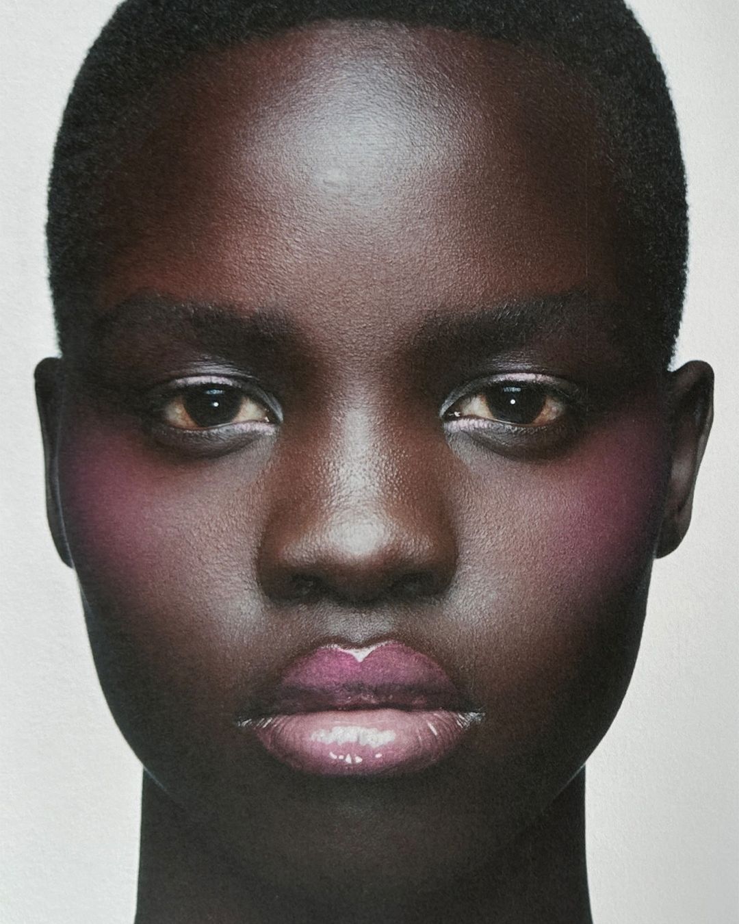 Close-up beauty portrait of a Black woman with short hair, glossy lips, and pink blush against a neutral background.