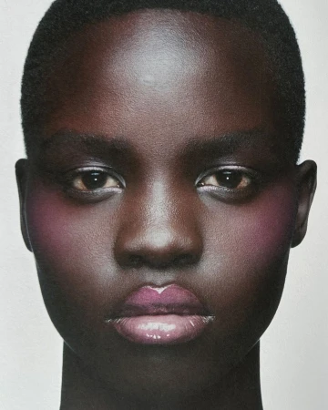 Close-up beauty portrait of a Black woman with short hair, glossy lips, and pink blush against a neutral background.