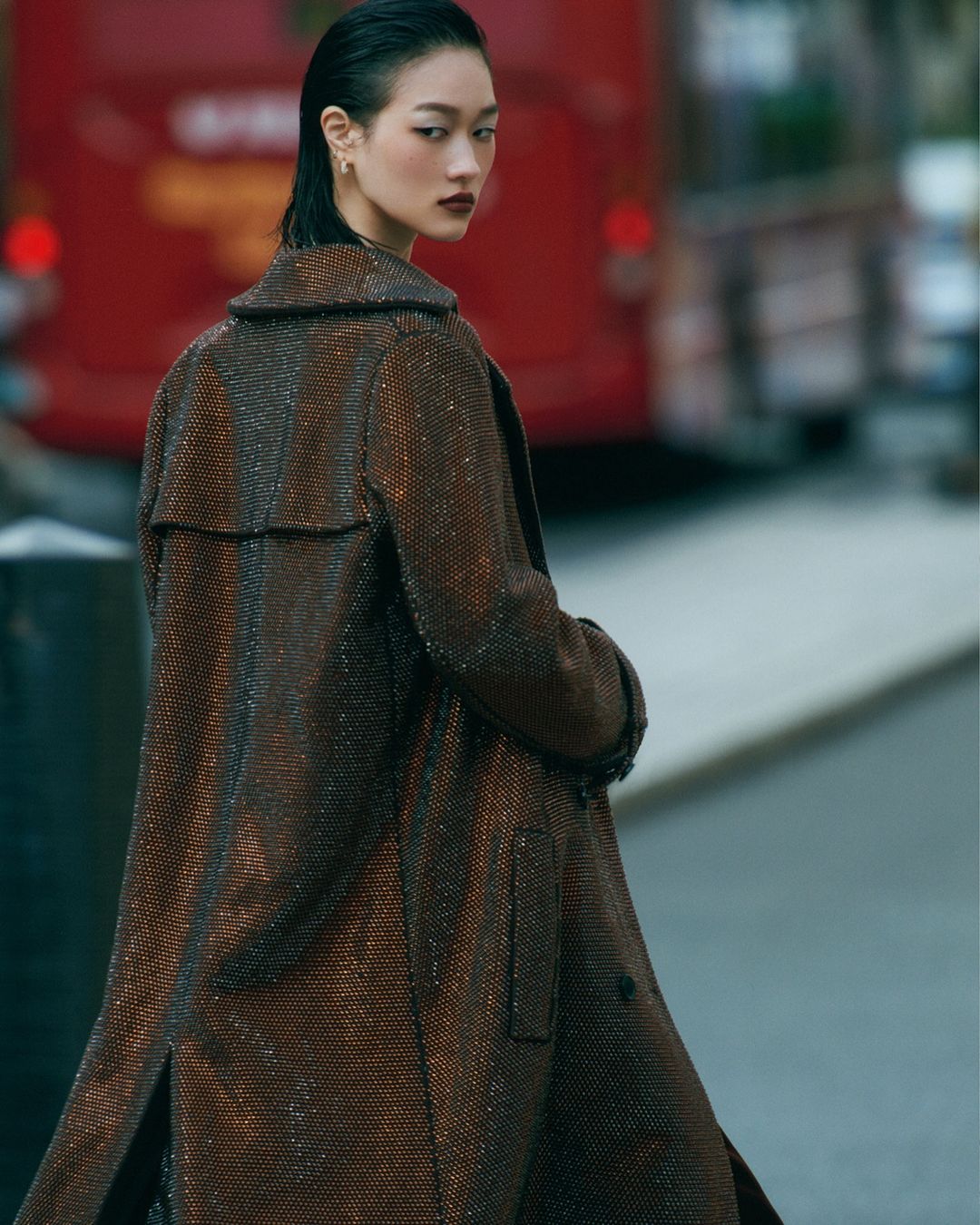Asian female fashion model in a brown patterned coat, looking over her shoulder on a city street with a red bus in the background.