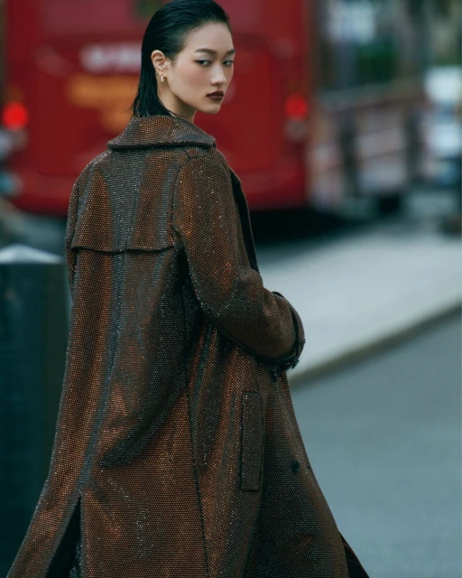 Asian female fashion model in a brown patterned coat, looking over her shoulder on a city street with a red bus in the background.