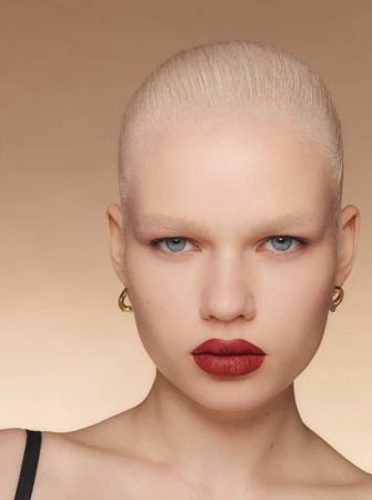 Close-up portrait of a model with short blonde hair and bold red lipstick wearing gold hoop earrings against a neutral gradient background.