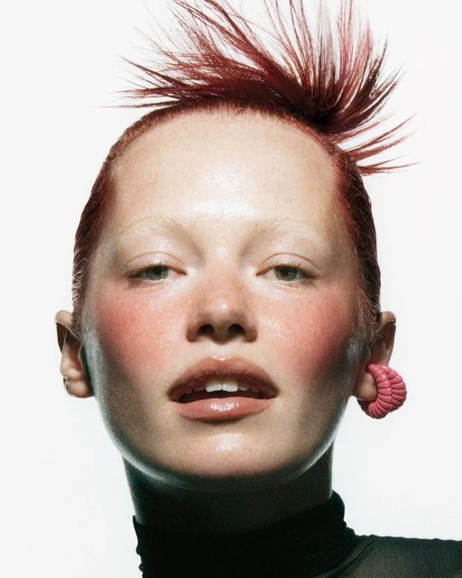 Close-up portrait of a young adult female with red spiky hair, bright pink makeup, and an earring, set against a white background.