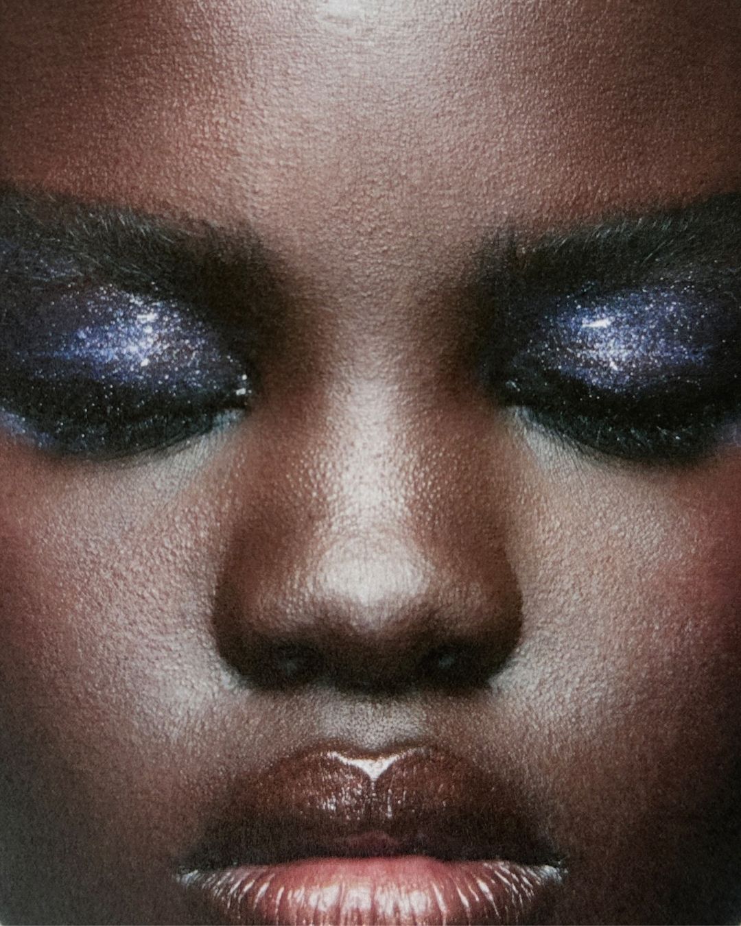 Close-up of an adult female face, dark skin, closed eyes, and glossy lips. The woman is wearing eye makeup with a glittering finish.