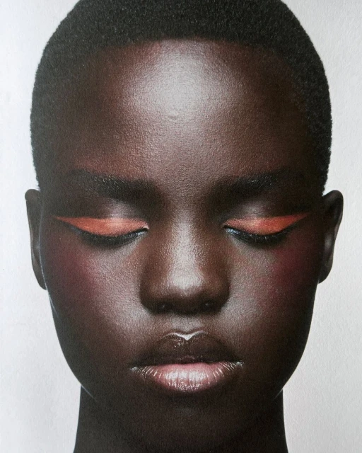 Close-up beauty shot of a woman with dark skin, closed eyes, orange eyeliner, and glossy lips, set against a bright background.