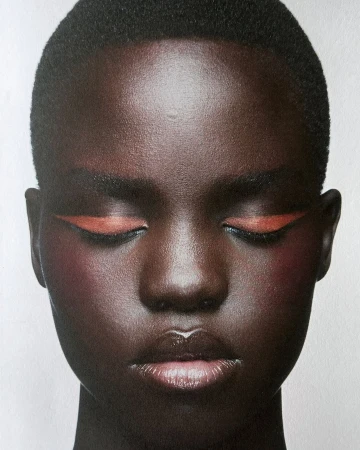 Close-up beauty shot of a woman with dark skin, closed eyes, orange eyeliner, and glossy lips, set against a bright background.