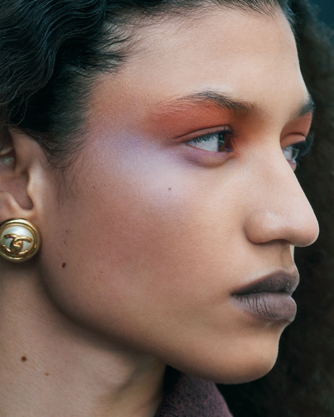 Close-up portrait of a woman with warm makeup, iridescent eye shadow, dark lipstick, and a vintage gold clip earring.