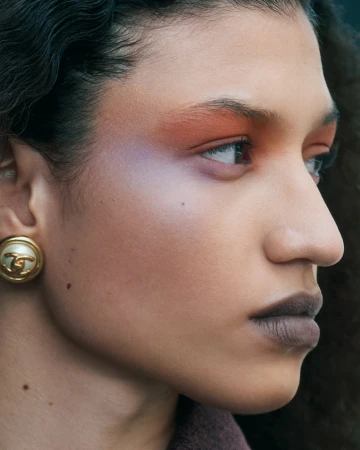 Close-up portrait of a woman with warm makeup, iridescent eye shadow, dark lipstick, and a vintage gold clip earring.