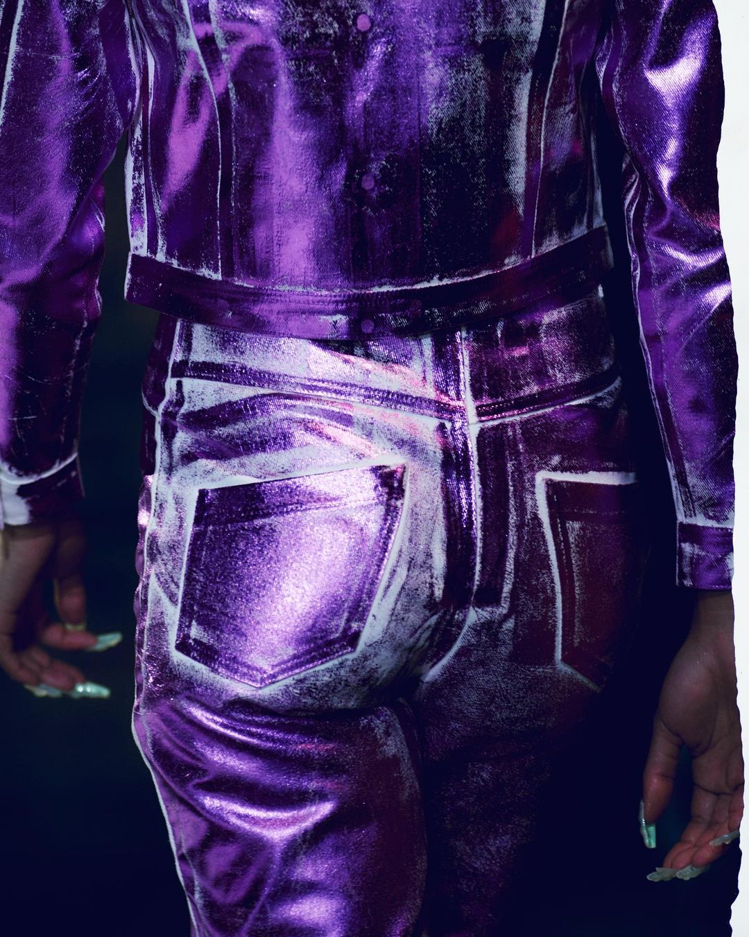 Back view of a person in a shiny, textured purple metallic jacket and jeans, with long green nails visible on their hands.