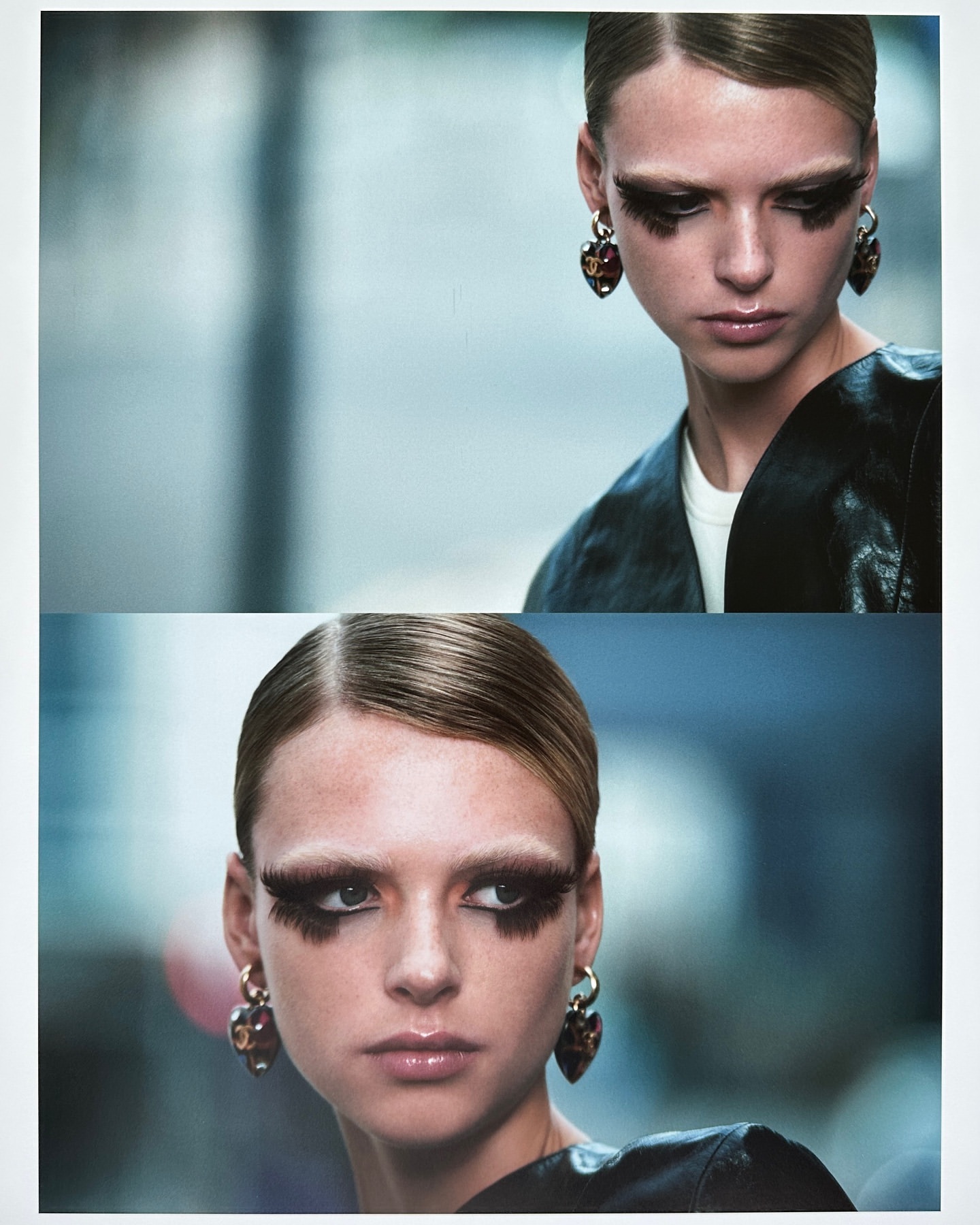 Close-up portrait of a woman in a black jacket and hoop earrings, featuring oversized eyelashes and a blurred blue-grey backdrop in the studio.