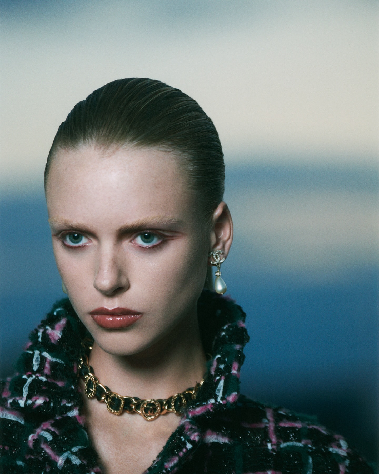 Close-up of a stylish adult female model with slicked-back hair wearing Chanel jewelry and a textured jacket against a blue gradient background.