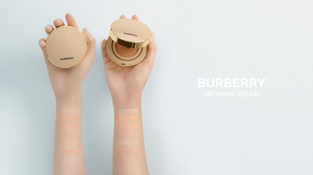 Cosmetic foundation compacts are displayed in two hands against a light blue background, with the brand Burberry and product name visible.