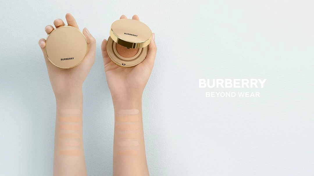 Cosmetic foundation compacts are displayed in two hands against a light blue background, with the brand Burberry and product name visible.
