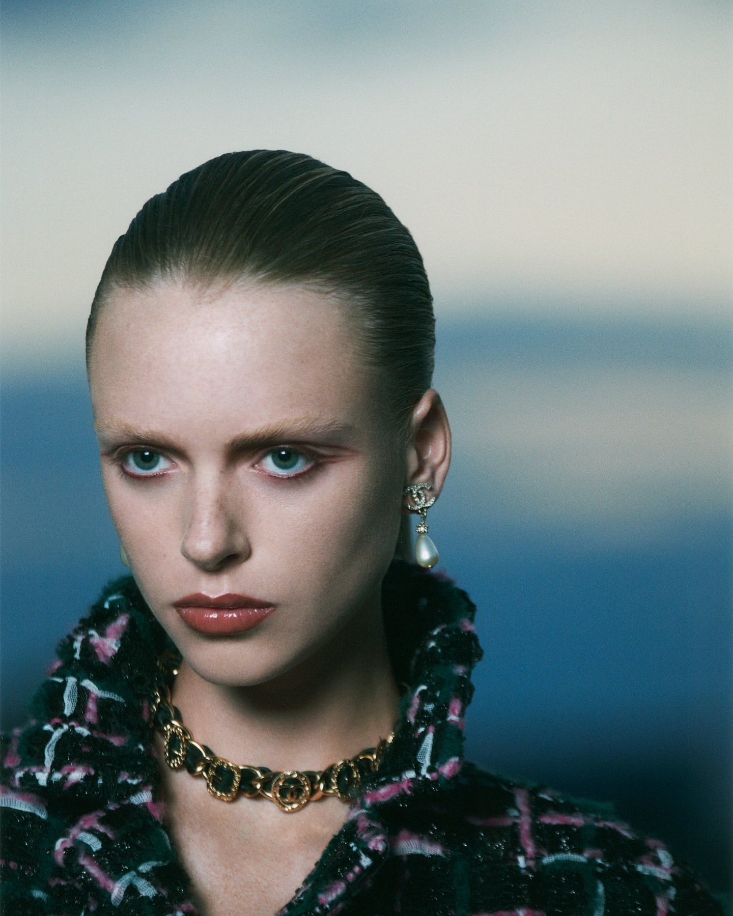 Close-up of a stylish adult female model with elegant jewelry, wearing a tweed jacket against a soft, blurred blue background.