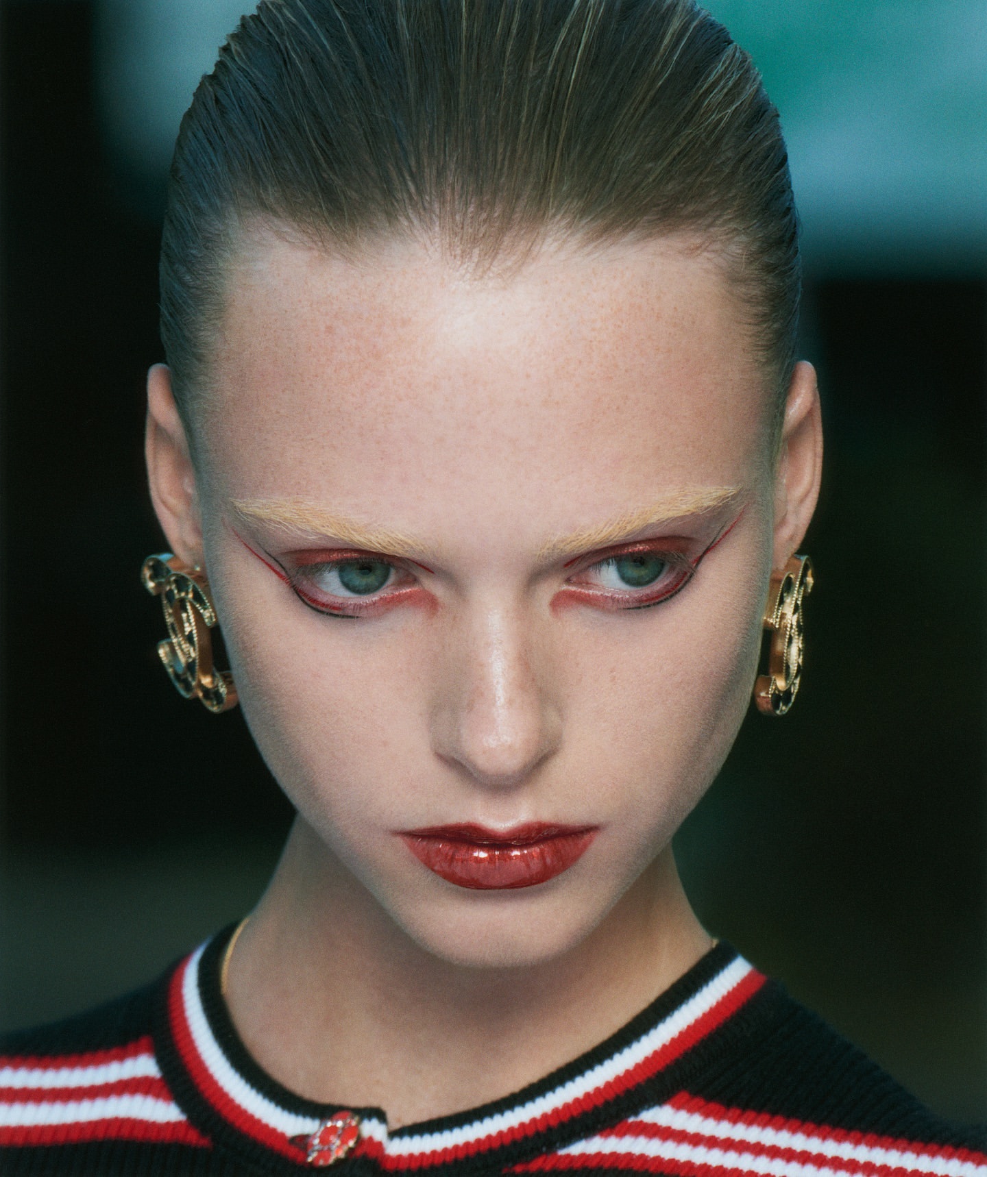 Close-up portrait of a woman with slicked-back hair, statement gold earrings, and dramatic red eyeliner and lipstick, fashion beauty shot.