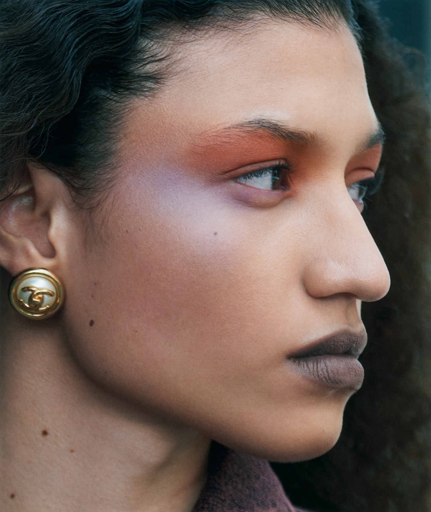 Close-up of a woman with curly dark hair, wearing statement earrings and wearing eye makeup looking slightly up and off to the side.