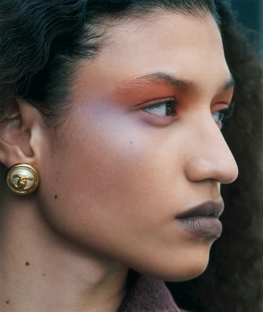 Close-up of a woman with curly dark hair, wearing statement earrings and wearing eye makeup looking slightly up and off to the side.