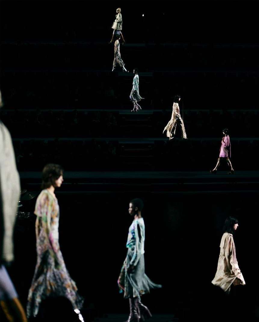 Fashion show on a dark stage with models walking in flowing dresses, illuminated by spotlights creating an ethereal and elegant atmosphere.