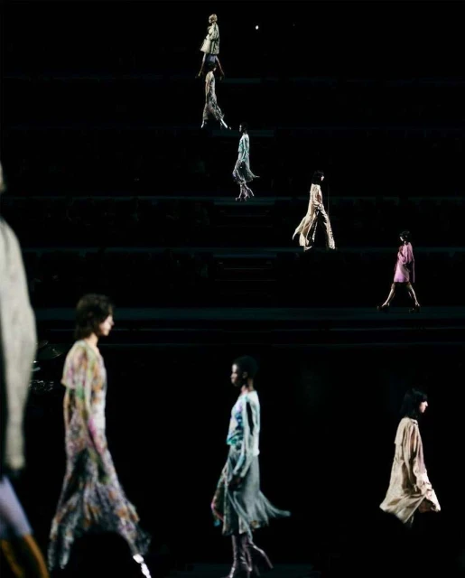 Fashion show on a dark stage with models walking in flowing dresses, illuminated by spotlights creating an ethereal and elegant atmosphere.