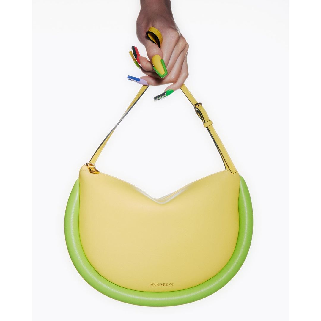 Close-up of a hand holding a yellow leather JW Anderson handbag with green piping, set against a stark white background.