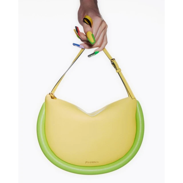 Close-up of a hand holding a yellow leather JW Anderson handbag with green piping, set against a stark white background.