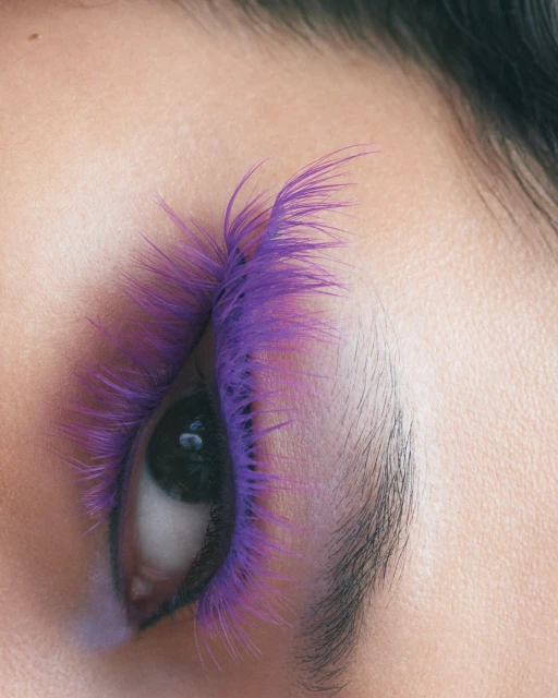 Close-up of a woman's eye with purple feathered false eyelashes, highlighted by soft lighting against a light skin tone.