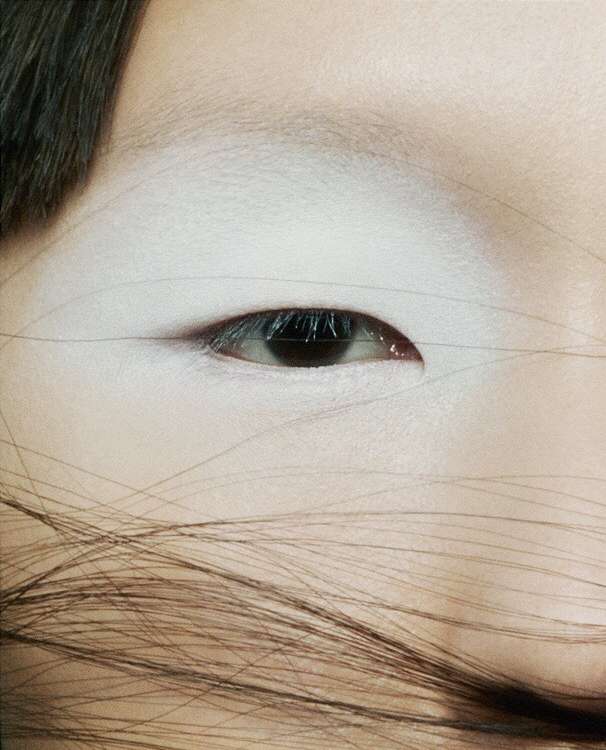 Close-up of a woman's face showing an eye with dark iris, surrounded by skin with white makeup, and brown hair across the lower face.