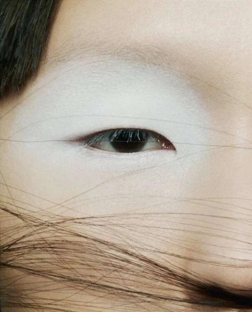 Close-up of a woman's face showing an eye with dark iris, surrounded by skin with white makeup, and brown hair across the lower face.